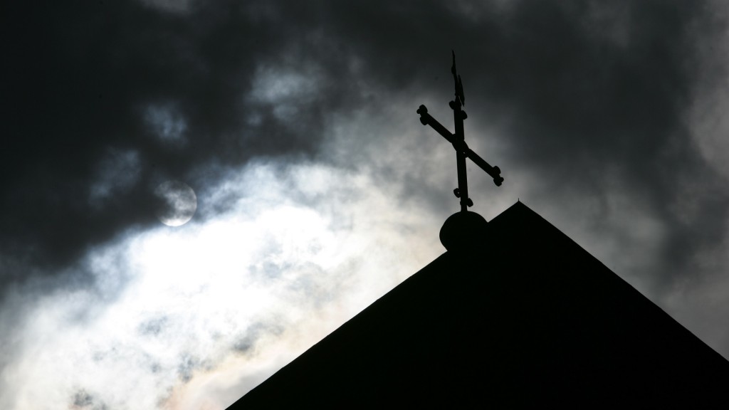 Im Gegenlicht und vor wolkenverhangenem Himmel ist die Kirchturmspitze mit Kreuz zu sehen. 
