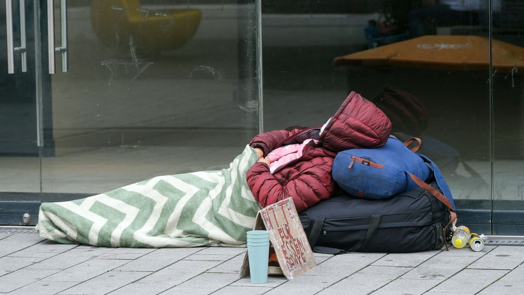 Eine obdachlose Person liegt mit Jacke und Decke vor einem leerstehenden Ladenlokal auf dem Gehweg