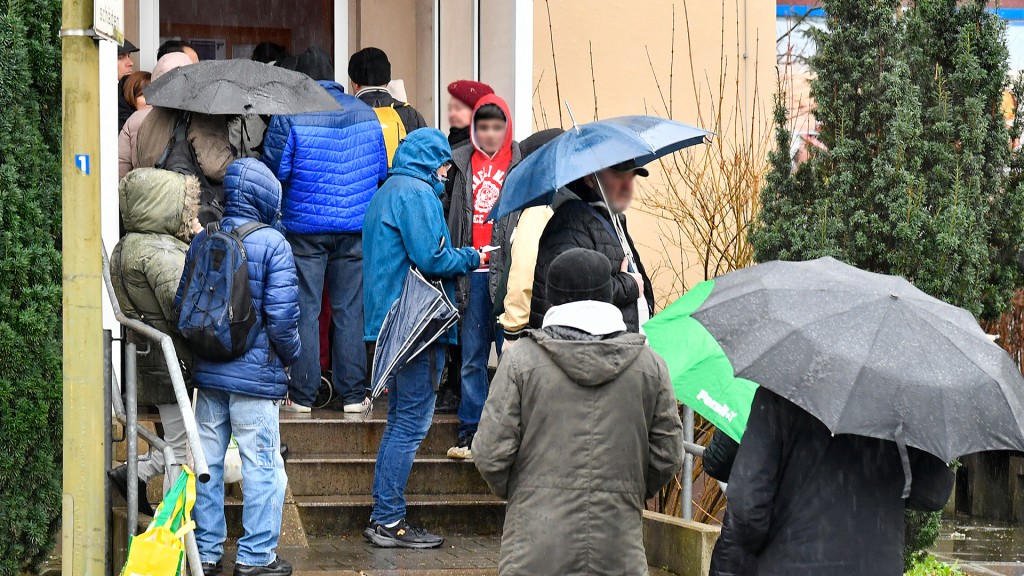 Foto: Menschen stehen mit Jacken und Schirmen vor einem Gebäude