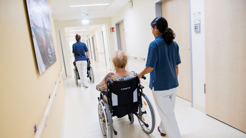 Zwei Pflegerinnen schieben Patienten mit Rollstühlen durch einen Flur
