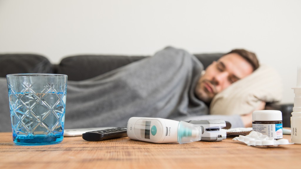 Ein Mann liegt krank auf einem Sofa, vor ihm auf dem Tisch Medikamente und ein Thermometer.