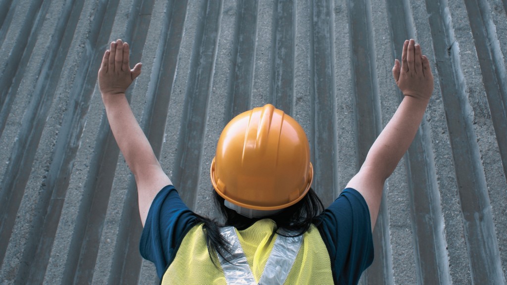 Das Bild oben zeigt eine Frau mit einem gelben Baustellenhelm, die vor einer grauen Betonwand steht. Sie hat die Arme erhoben. 