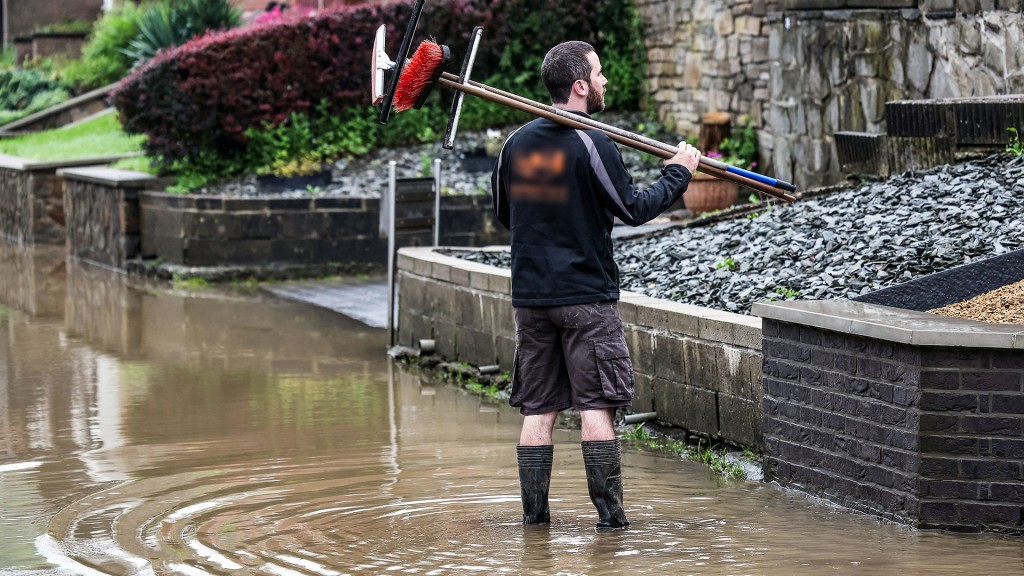 Ein Mann steht mit Gummistiefeln, Besen und Abziehern im Hochwasser.