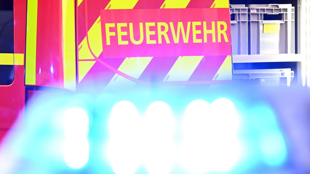 Ein Fahrzeug der Feuerwehr steht bei einem Einsatz auf einer Straße, während im Vordergrund das Blaulicht eines Polizeifahrzeugs zu sehen ist