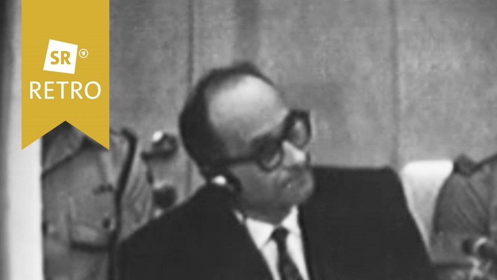 Adolf Eichmann im Gerichtssaal. Er trägt Kopfhörern und hat Akten vor sich ausgebreitet