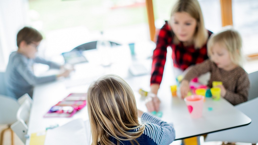Kinder sitzen am Tisch und werden von einer Lehrerin bei Aufgaben unterstützt.