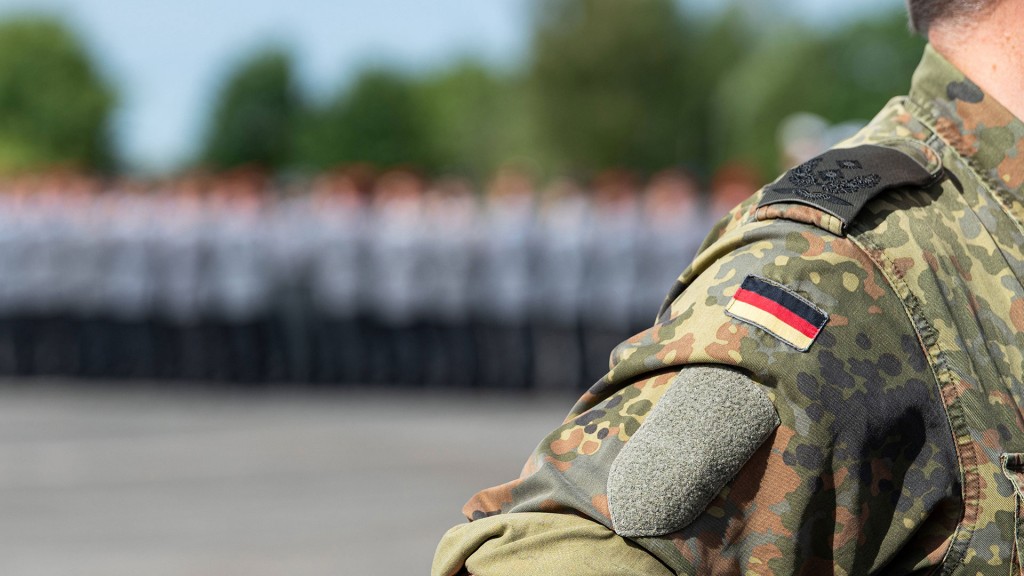Foto: Ein Soldat der Bundeswehr steht in Flecktarnuniform mit Deutschlandabzeichen auf dem Ärmel und eine Truppe im Hintergrund